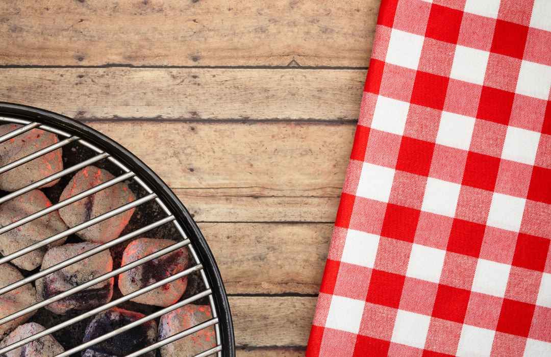 Grill with lit charcoal next to a red and white checkered tablecloth on a wooden surface.