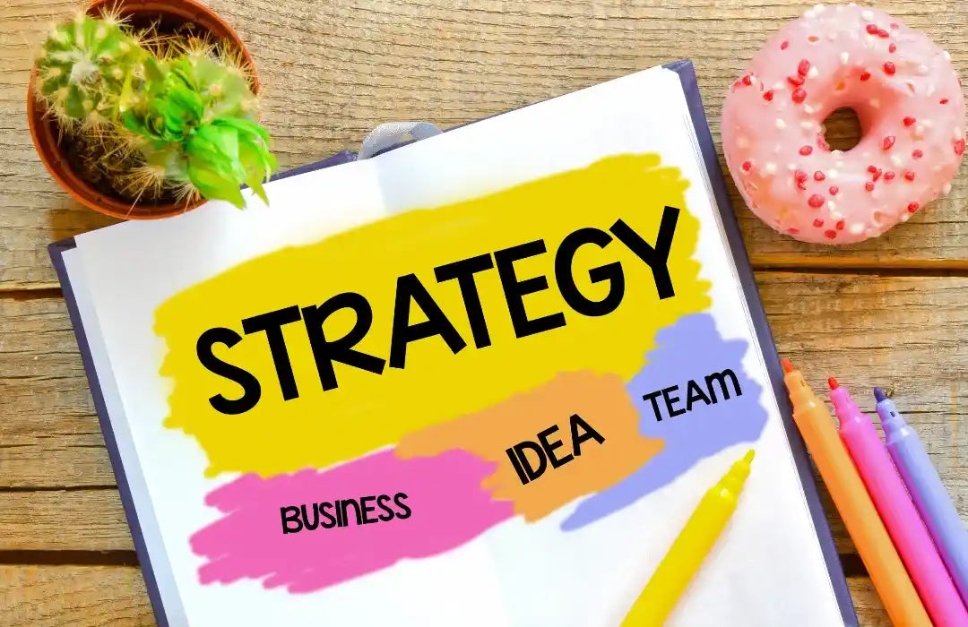 Notebook with the word “Strategy” highlighted, surrounded by notes labeled business, idea, and team, with colorful pens and a donut on the desk.