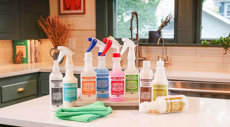 Medium closeup image of all nine liquid Speed Cleaning household cleaners in a row on a kitchen counter with two green cleaning cloths nearby.