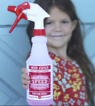 Photo of a little girl holding up a bottle of Red Juice Cleaner. She is about six years old, Caucaasian, with brown eyes and brown hair in a red blouse with yellow and white flowers. She is standing in front of a blue wood door.