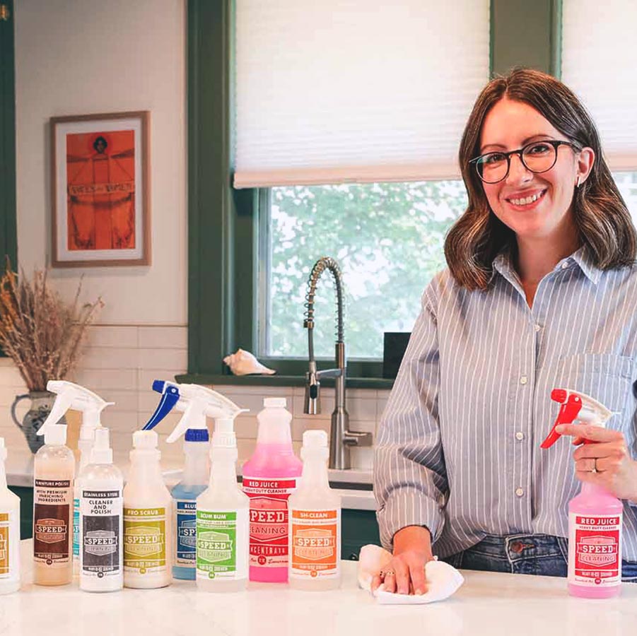 Closeup of the Speed Cleaning product line shown on a kitchen counter next to founder, Dr. Amy Sardone, PhD, who is holding a bottle of Red Juice in one hand and a cleaning cloth in the other.