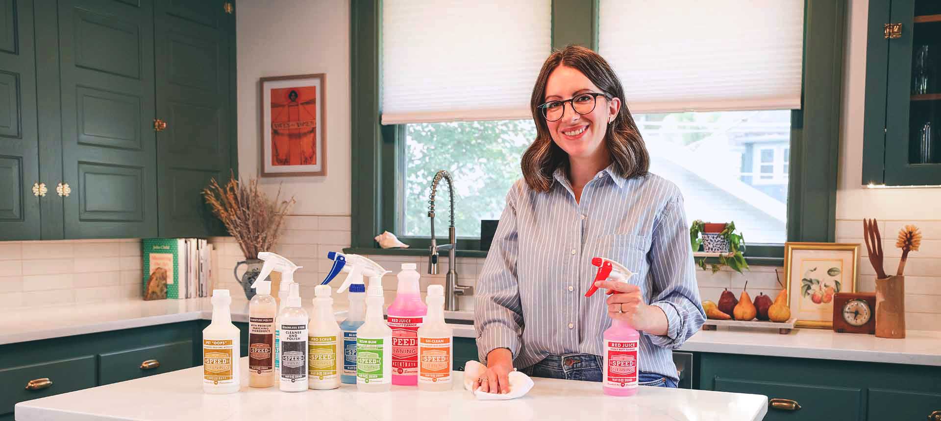 Speed Cleaning product line shown on a kitchen counter next to founder, Dr. Amy Sardone, PhD, who is holding a bottle of Red Juice in one hand and a cleaning cloth in the other.